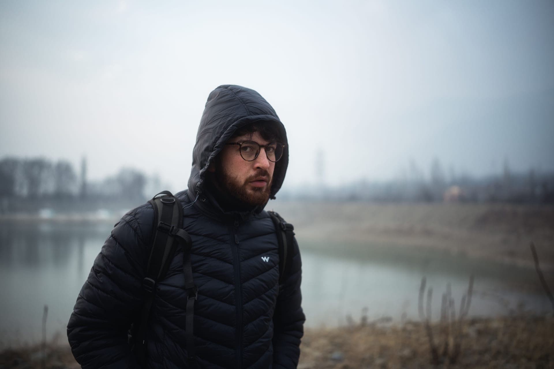 A man in glasses and a winter jacket beside a foggy lake
