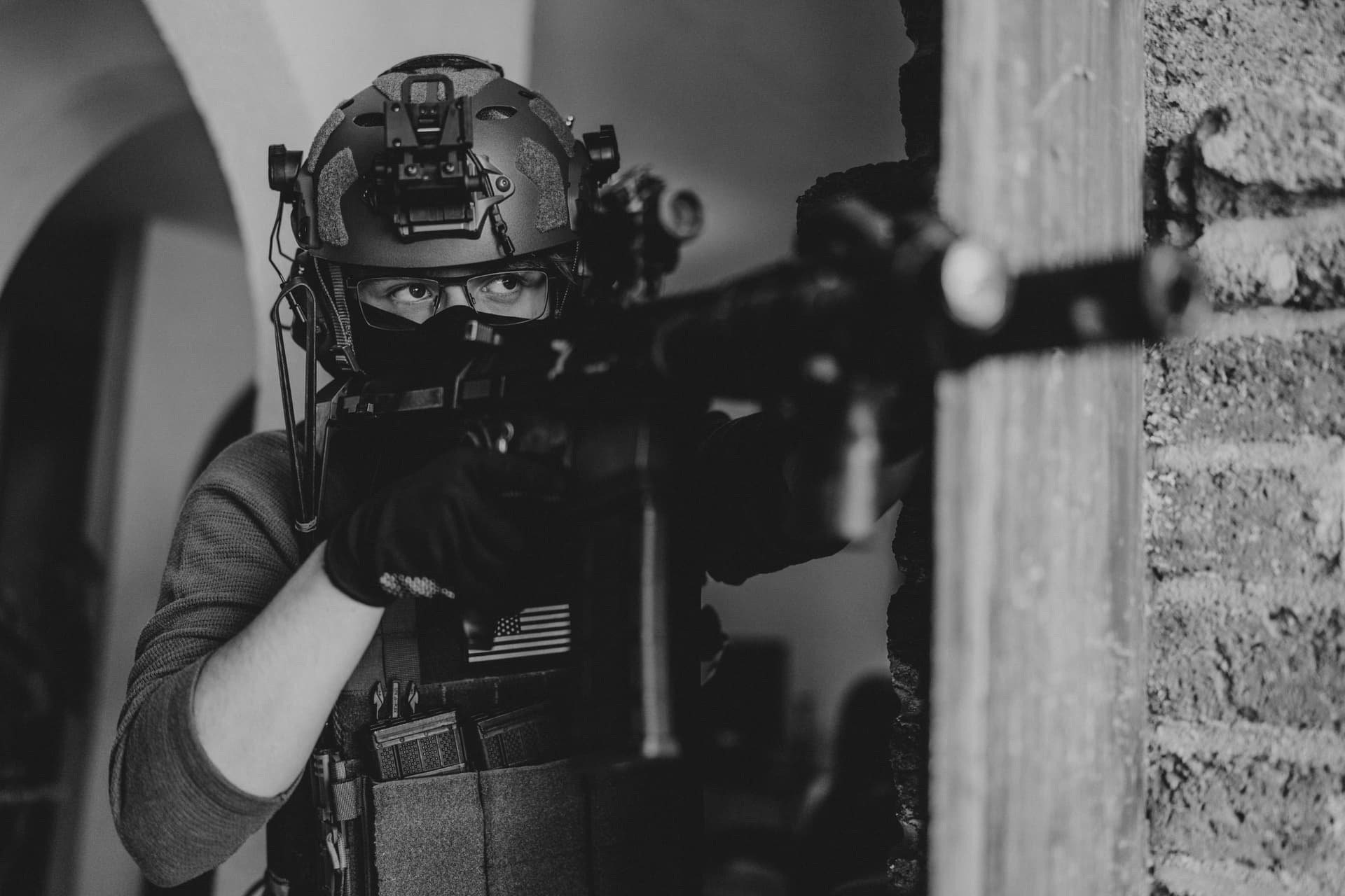 A soldier in tactical gear aiming a weapon indoors, in black and white