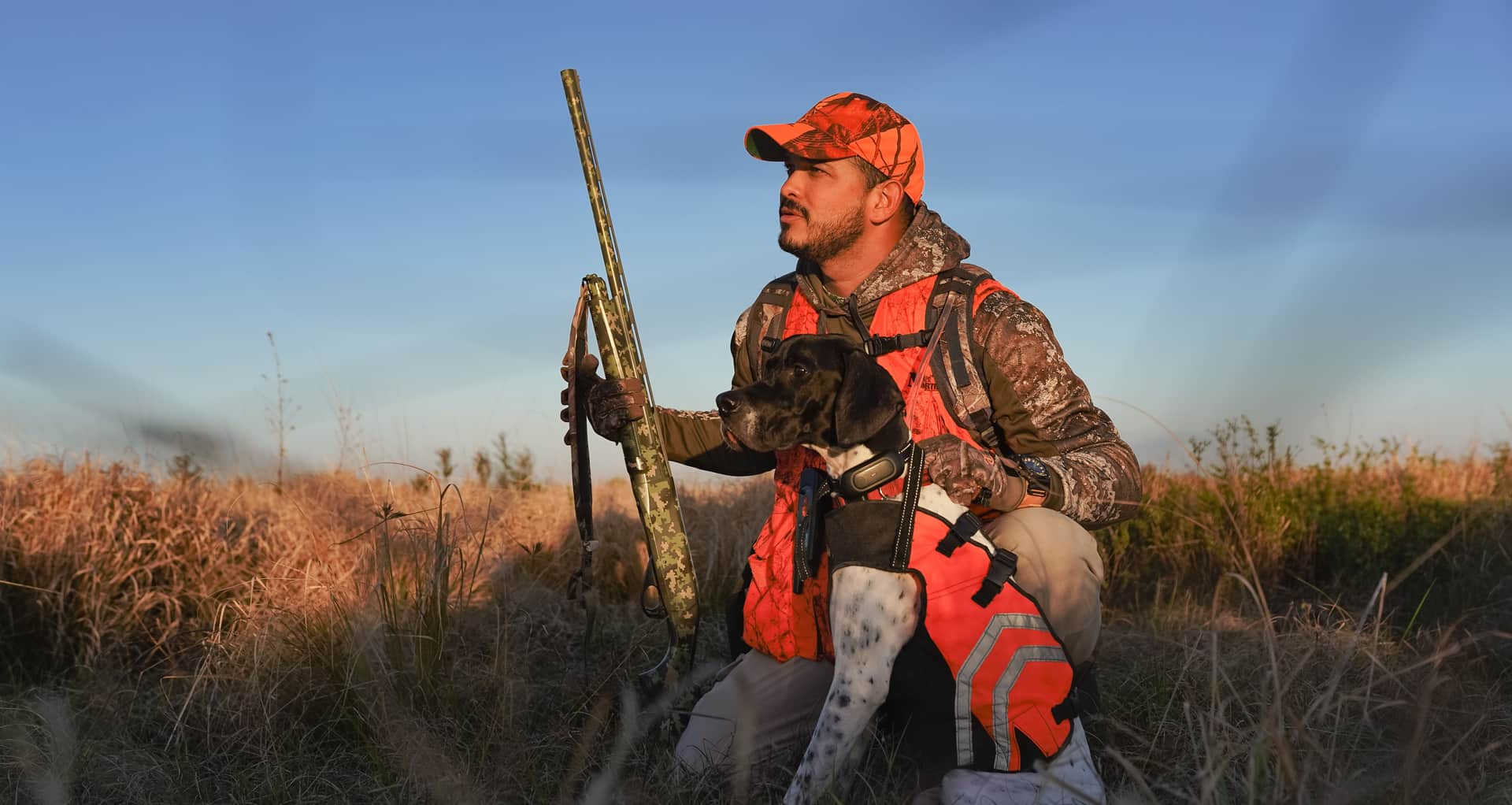 A hunter in camouflage with a dog and rifle in a meadow at sunrise
